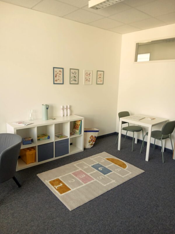 Ecke im Raum von Care Together mit einem REgal mit Spielsachen und Büchern, Bildern an der Wand, einem Tisch mit zwei Stühlen und einem Teppich.